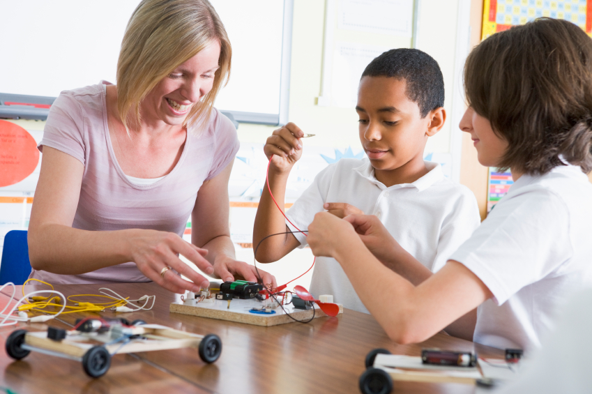 A teacher guiding two students in a hands-on STEM activity involving circuits and small vehicles. The students, wearing white uniforms, are engaged and working collaboratively on the project, while the teacher smiles and offers assistance. The scene is set in a brightly lit classroom with educational posters in the background.